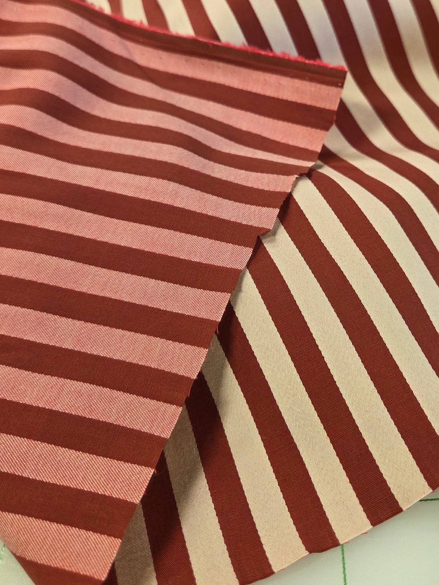Striped fabric with alternating dark red and cream vertical stripes, folded to show its texture and pattern, resting on a green grid cutting mat.