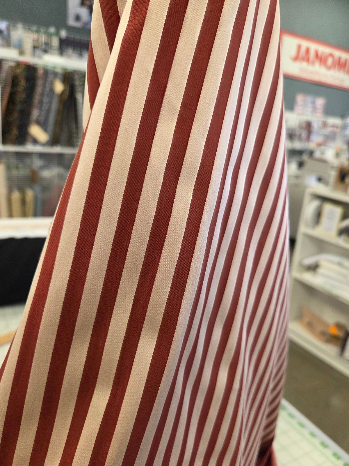 A close-up of a striped fabric, likely a scarf or sash, featuring alternating vertical stripes of deep red and cream. The fabric is draped in the foreground, with a blurred store background showing shelves and a sign that reads 'JANOME'.