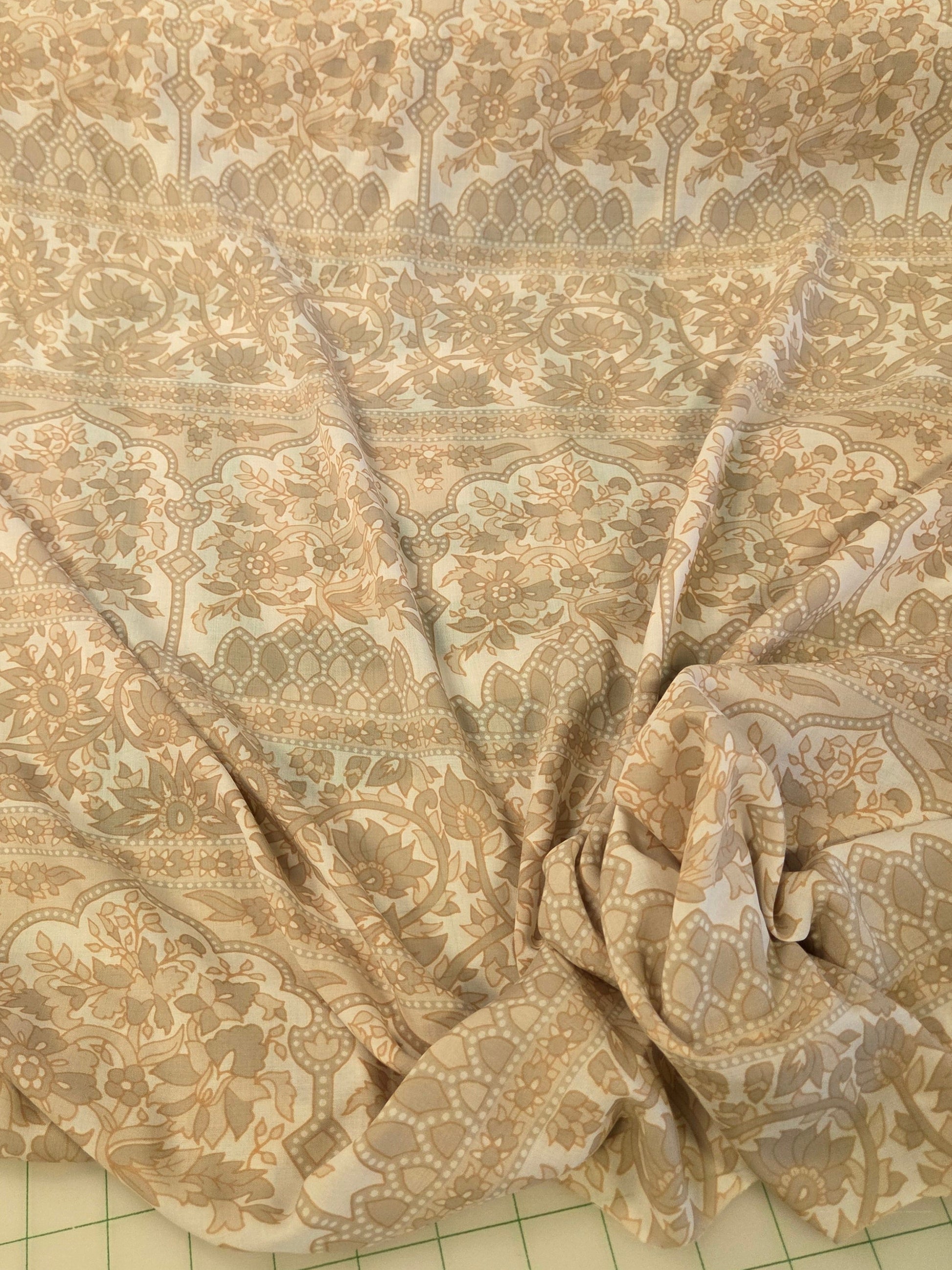 A beige and white patterned fabric, draped and gathered on a green grid cutting mat with a wooden ruler visible at the top.