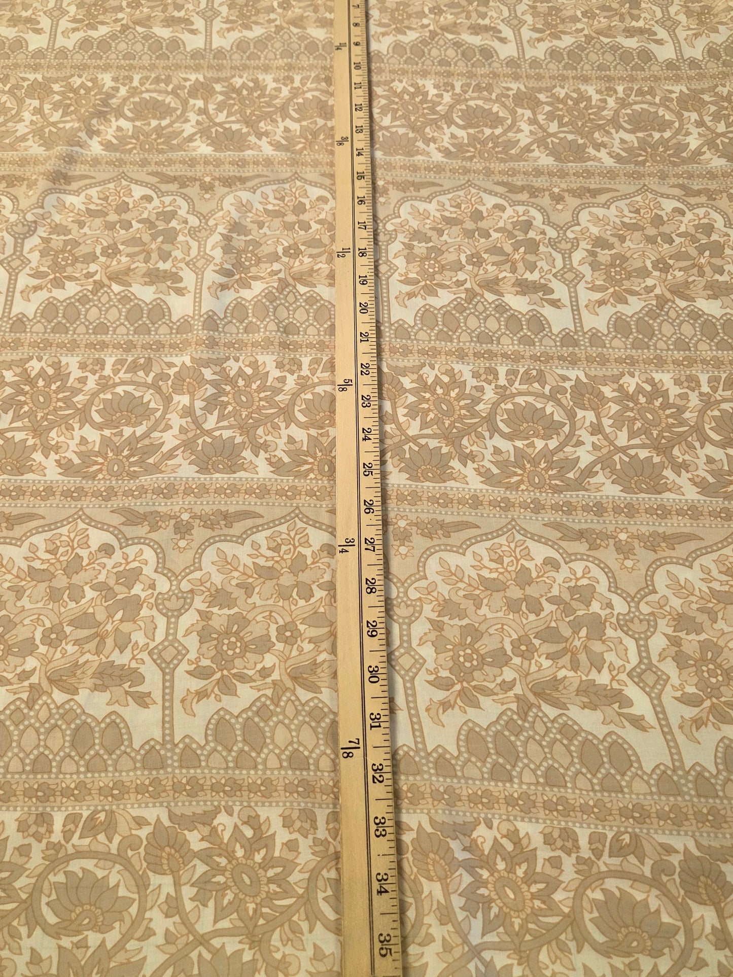A square piece of beige and white floral lace fabric laid flat on a green cutting mat, with a wooden ruler standing vertically in the center to show the pattern and scale.