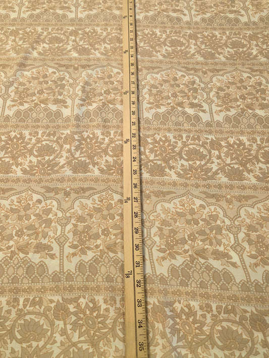 A square piece of beige and white floral lace fabric laid flat on a green cutting mat, with a wooden ruler standing vertically in the center to show the pattern and scale.
