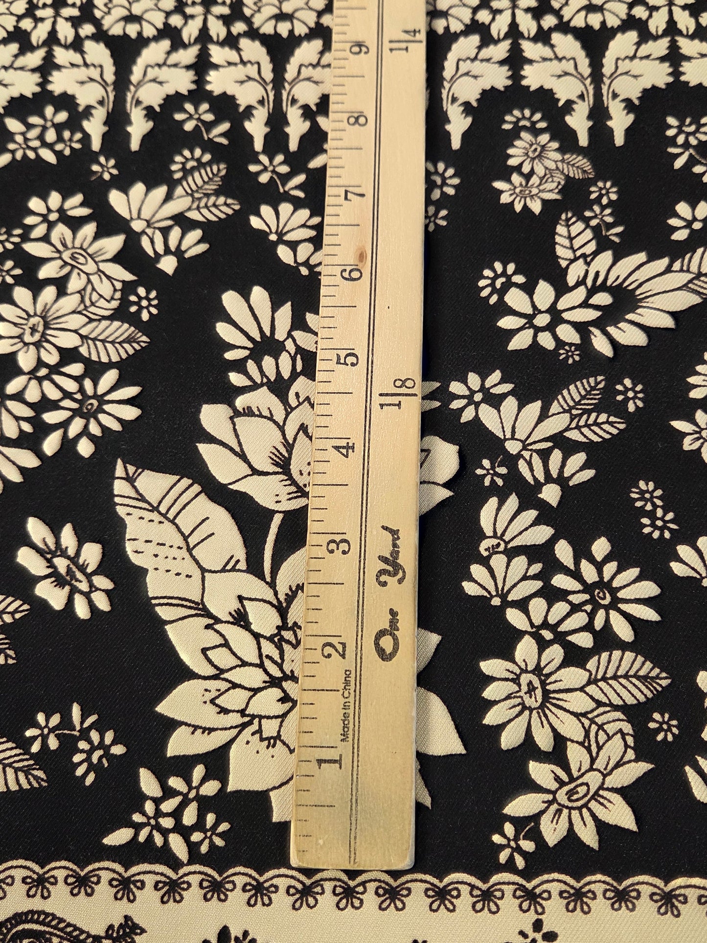Black and cream floral fabric with a paisley border, shown with a wooden yardstick ruler to indicate scale.