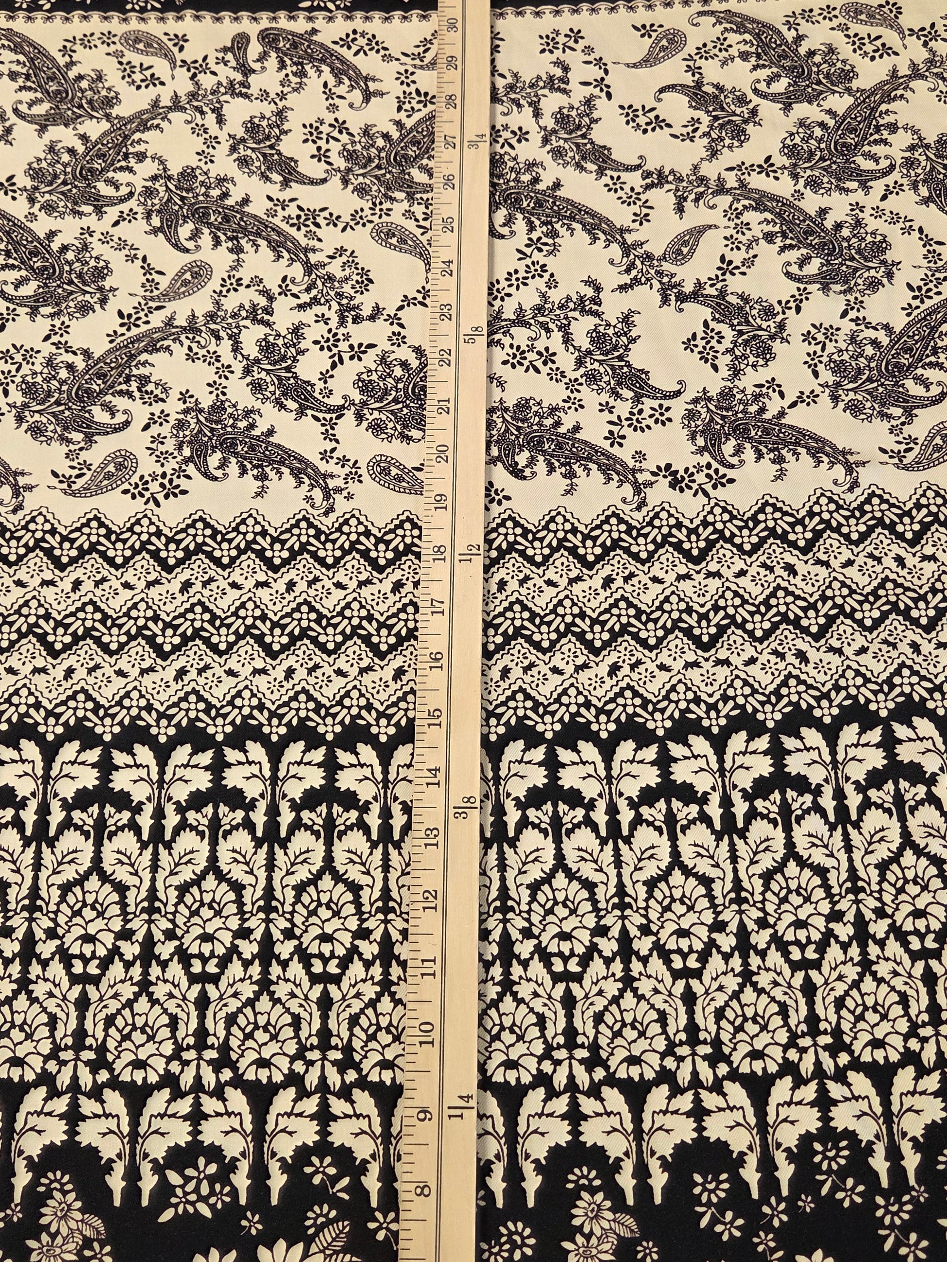 A close-up of a black and cream floral and paisley patterned fabric with a vertical ruler in the center for scale.