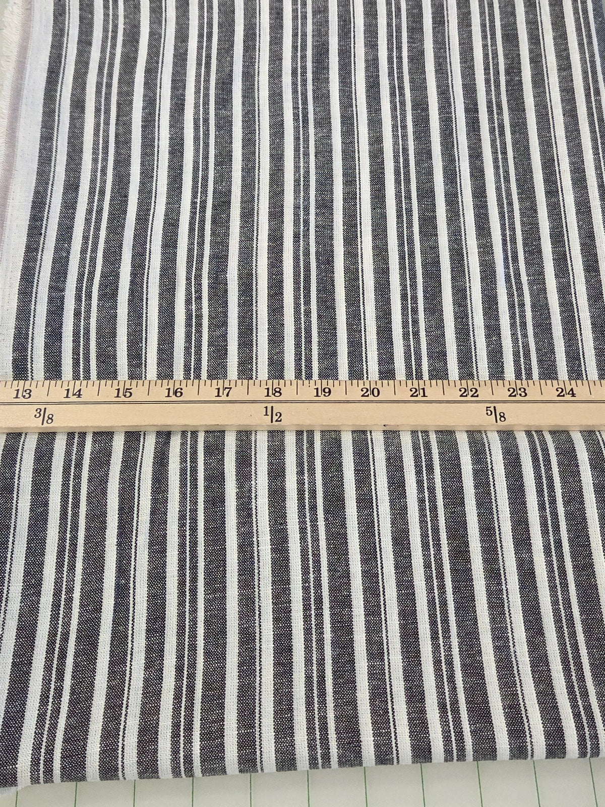 A folded piece of fabric with vertical black, gray, and white stripes, shown on a green grid cutting mat with a wooden ruler laid across the middle to indicate size.