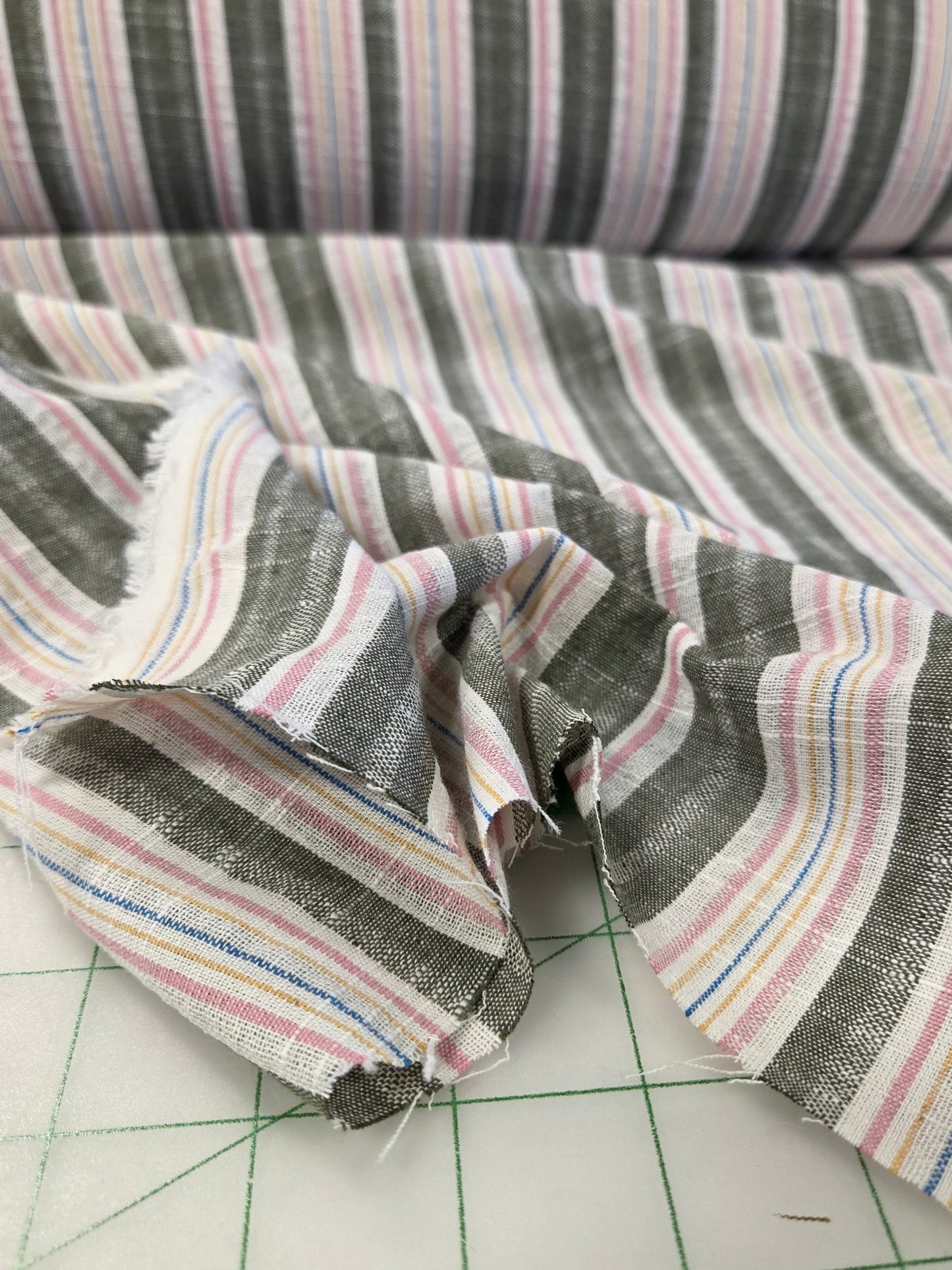 A close-up of striped fabric draped over a cutting mat. The fabric has a dark gray base with vertical stripes in pink, white, blue, and yellow.