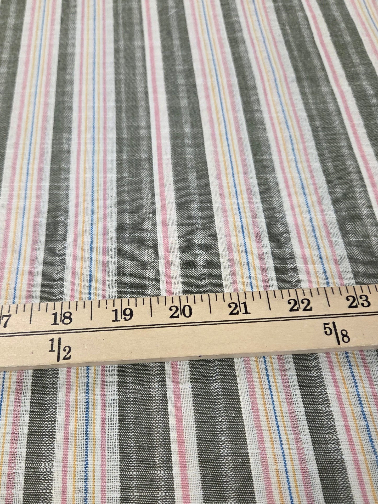 A close-up of striped fabric with vertical bands in gray, white, pink, blue, and yellow, with a wooden ruler laid horizontally to show scale.