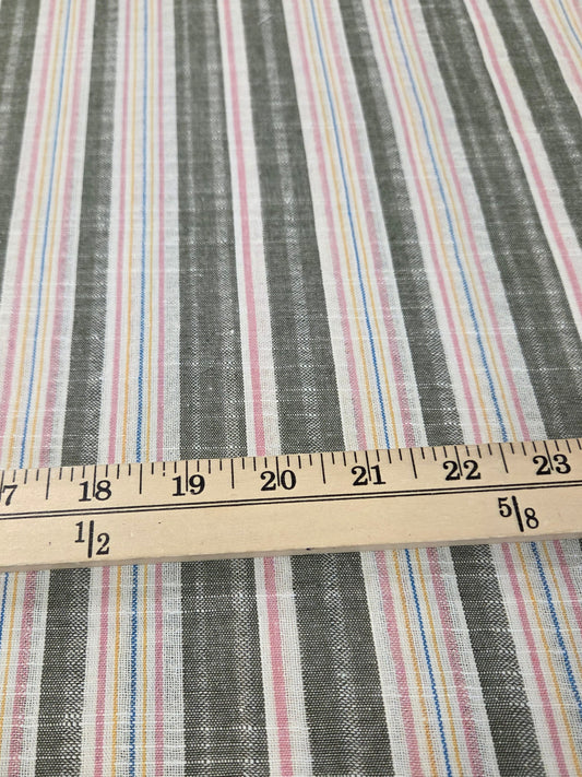 A close-up of striped fabric with vertical bands in gray, white, pink, blue, and yellow, with a wooden ruler laid horizontally to show scale.
