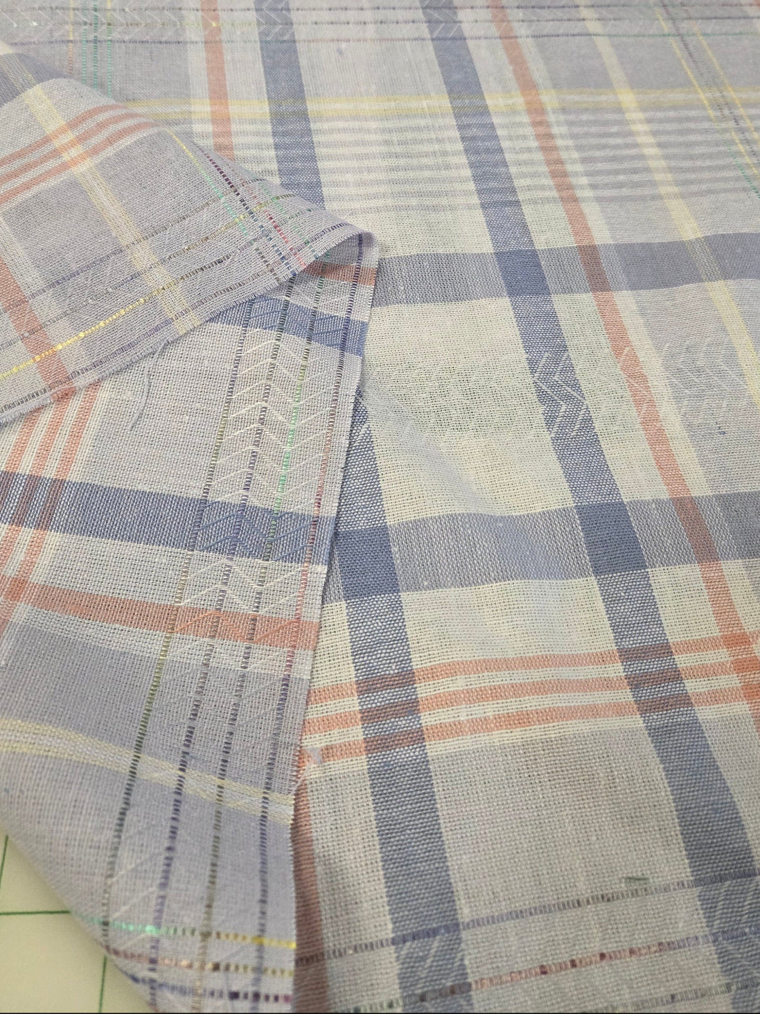 A close-up of plaid fabric with a light blue-gray base, white stripes, and thin orange and yellow lines, folded over to show the texture and stitching.