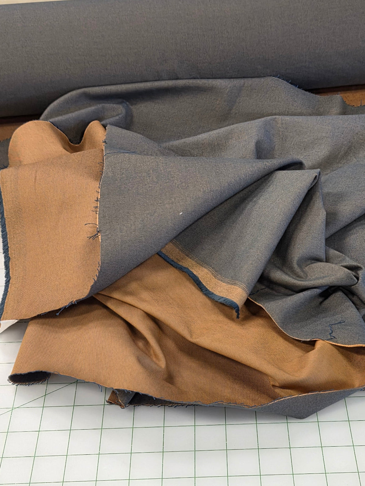 A pile of fabric pieces in gray and tan/brown, with a rolled piece of gray fabric visible at the top and a green grid cutting mat underneath.