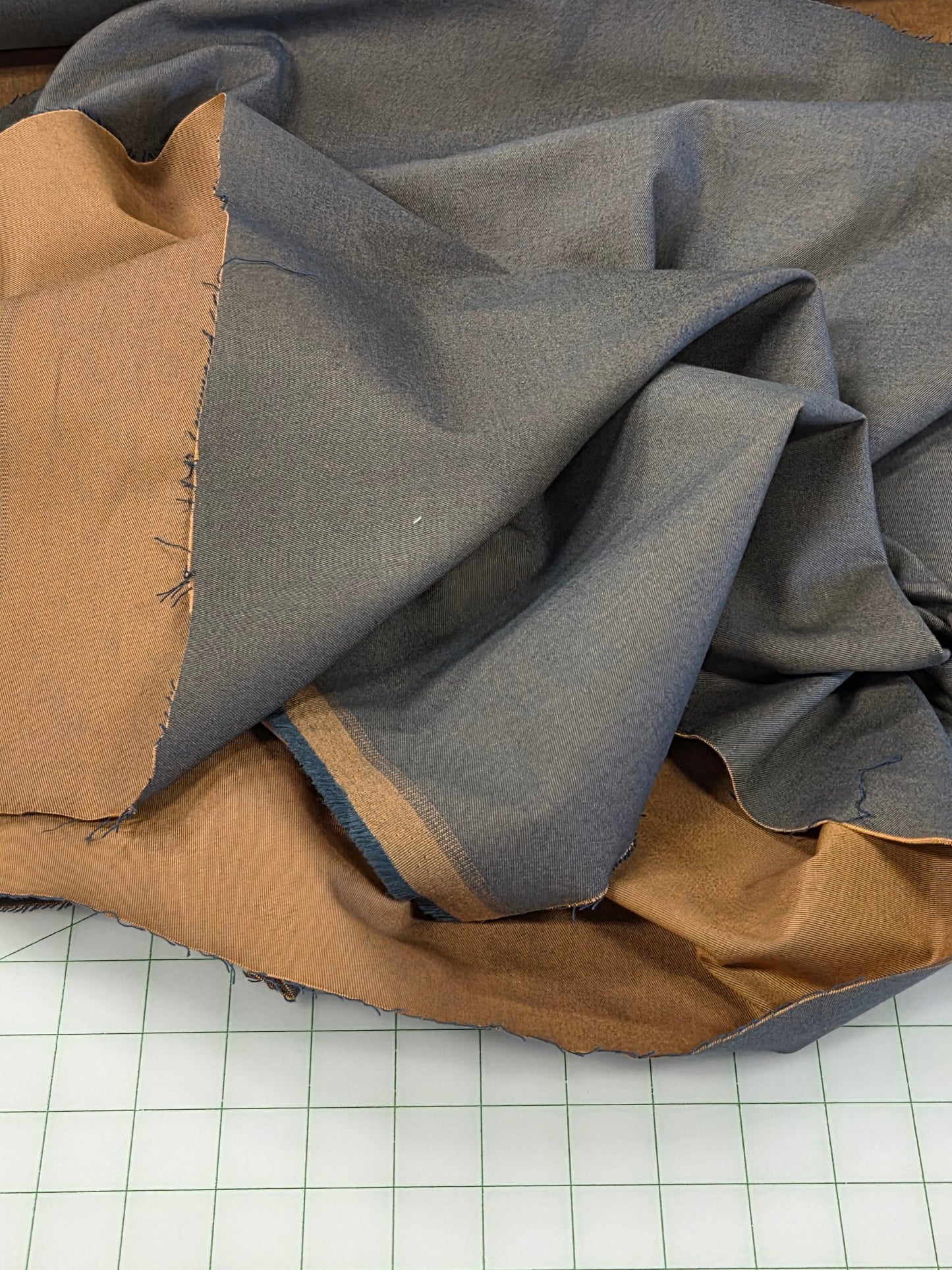 A piece of gray fabric with a tan lining is draped over a white grid cutting mat, with a roll of striped fabric visible in the background.