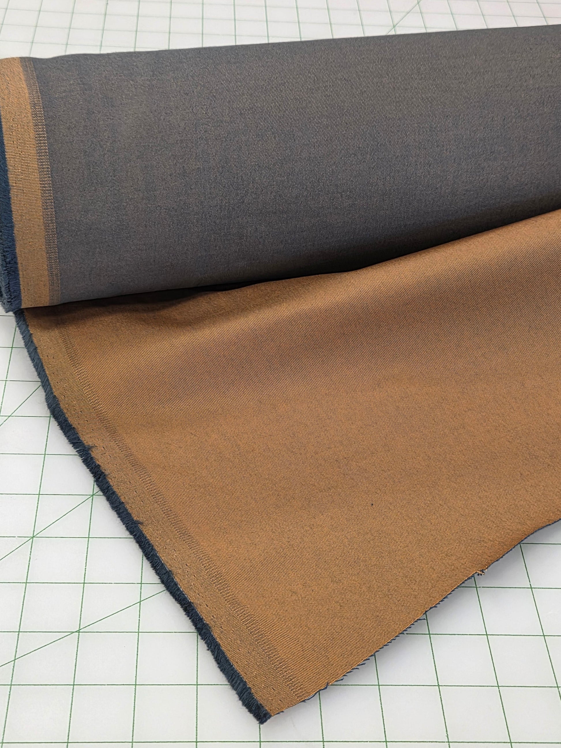 A roll of fabric with a dark gray top side and a warm brown/tan reverse side, laid on a white gridded cutting mat.