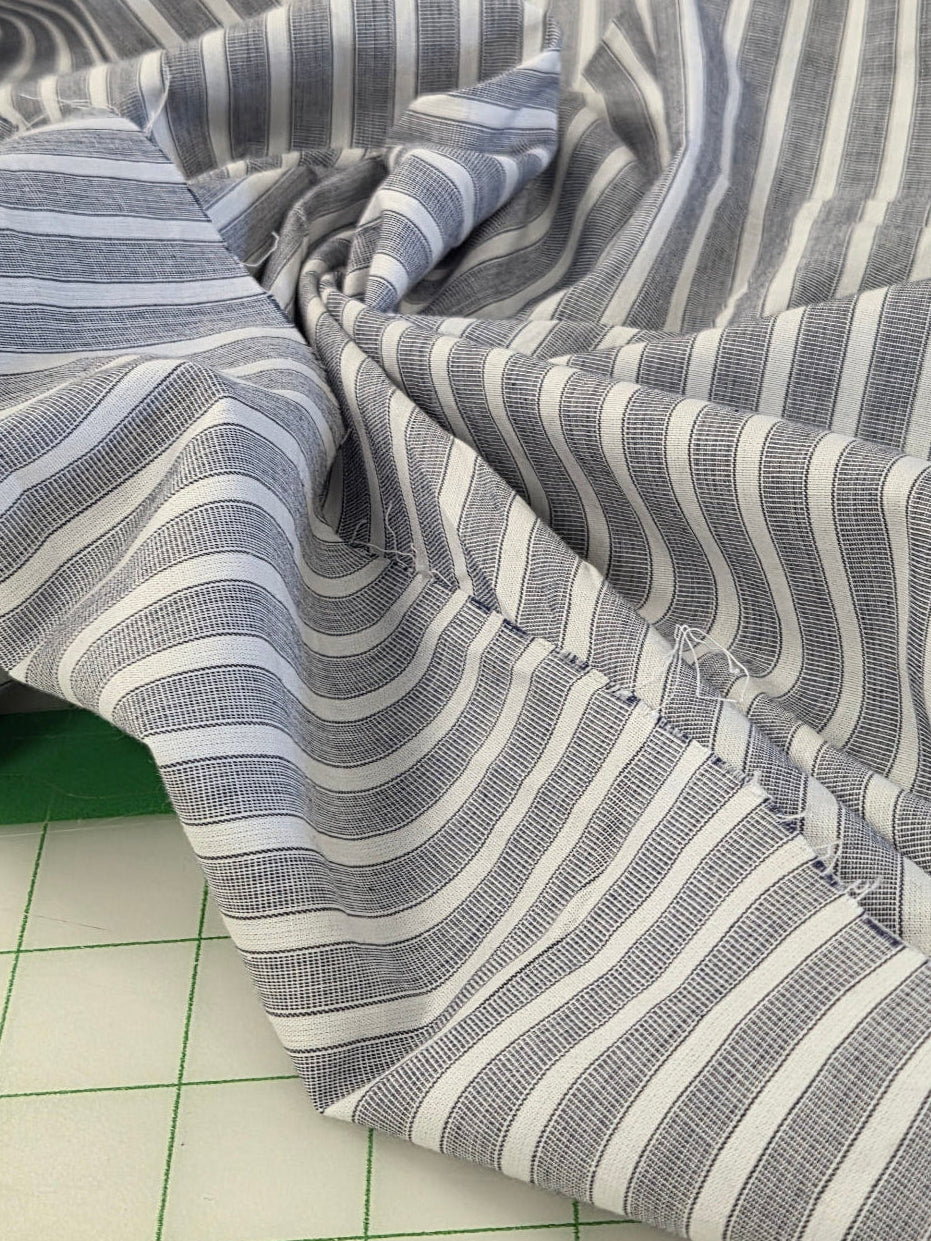 A close-up of draped striped fabric with alternating gray and white vertical stripes, shown gathered in the center over a green cutting mat and white grid surface.