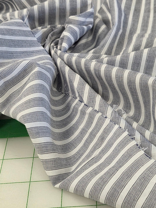 A close-up of draped striped fabric with alternating gray and white vertical stripes, shown gathered in the center over a green cutting mat and white grid surface.