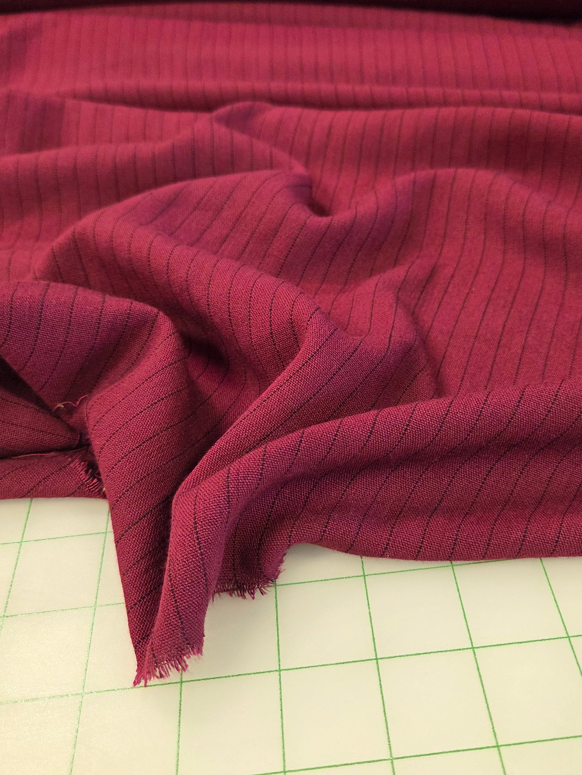 A close-up of draped burgundy fabric with a fine vertical stripe pattern, laid over a white gridded cutting mat.