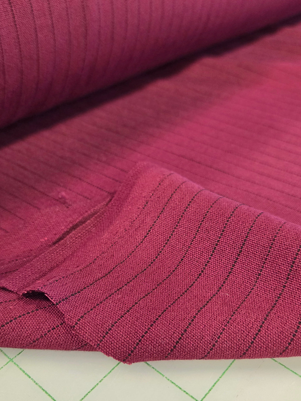 A close-up of folded magenta-pink fabric with thin black vertical pinstripes, resting on a white cutting mat with green grid lines.