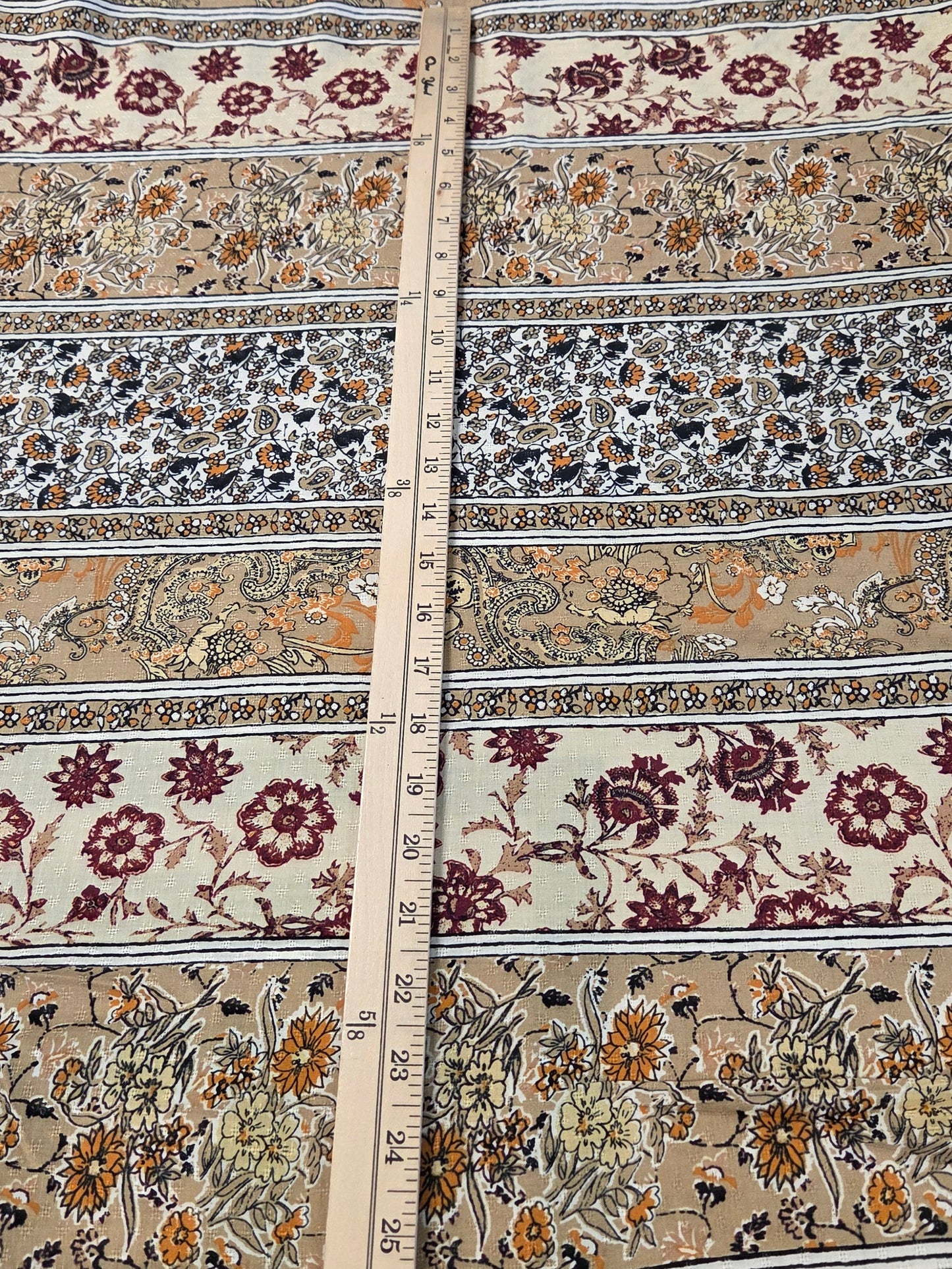 A close-up of a patterned fabric laid flat, showing horizontal bands of floral and ornamental motifs in beige, rust red, mustard yellow, gray, and black. A wooden ruler is placed vertically through the center to indicate scale.