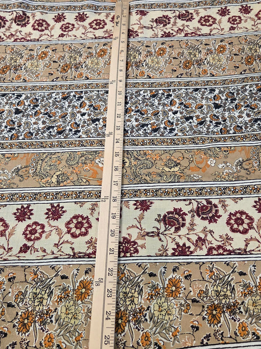 A close-up of a patterned fabric laid flat, showing horizontal bands of floral and ornamental motifs in beige, rust red, mustard yellow, gray, and black. A wooden ruler is placed vertically through the center to indicate scale.