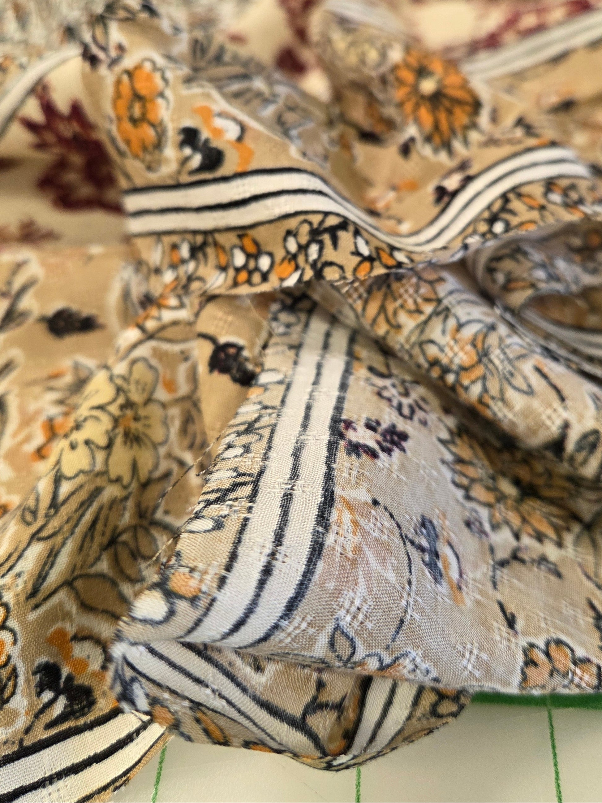 A close-up of a lightweight patterned fabric with a beige base, featuring orange floral motifs and black-and-white striped borders.