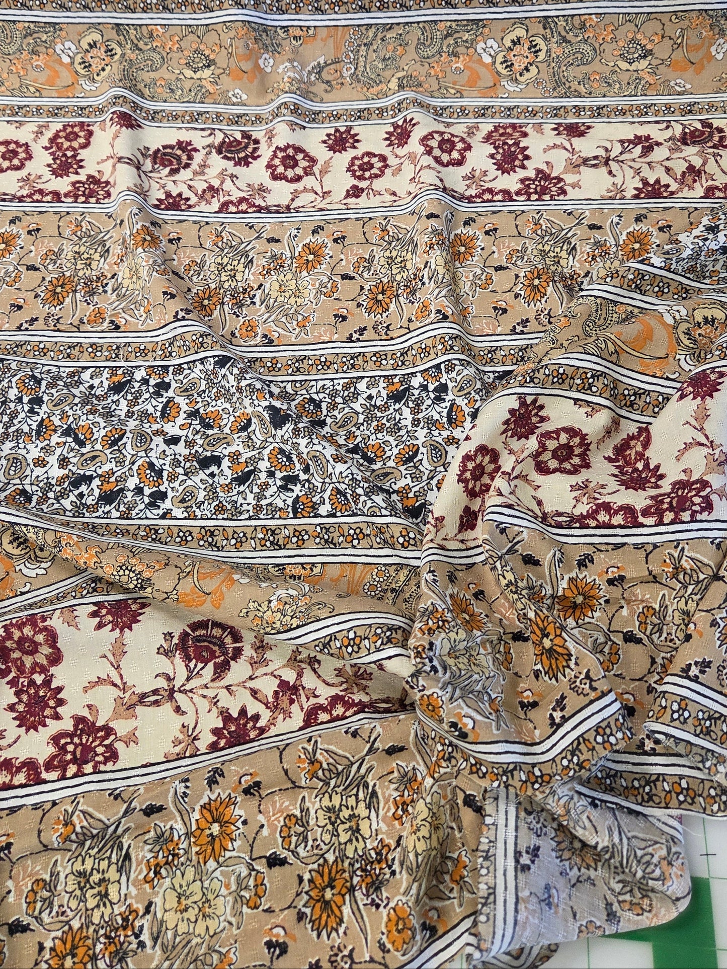 A draped piece of fabric with an intricate, multi-colored floral and ornamental pattern in beige, burgundy, orange, and gray, laid out on a green surface.