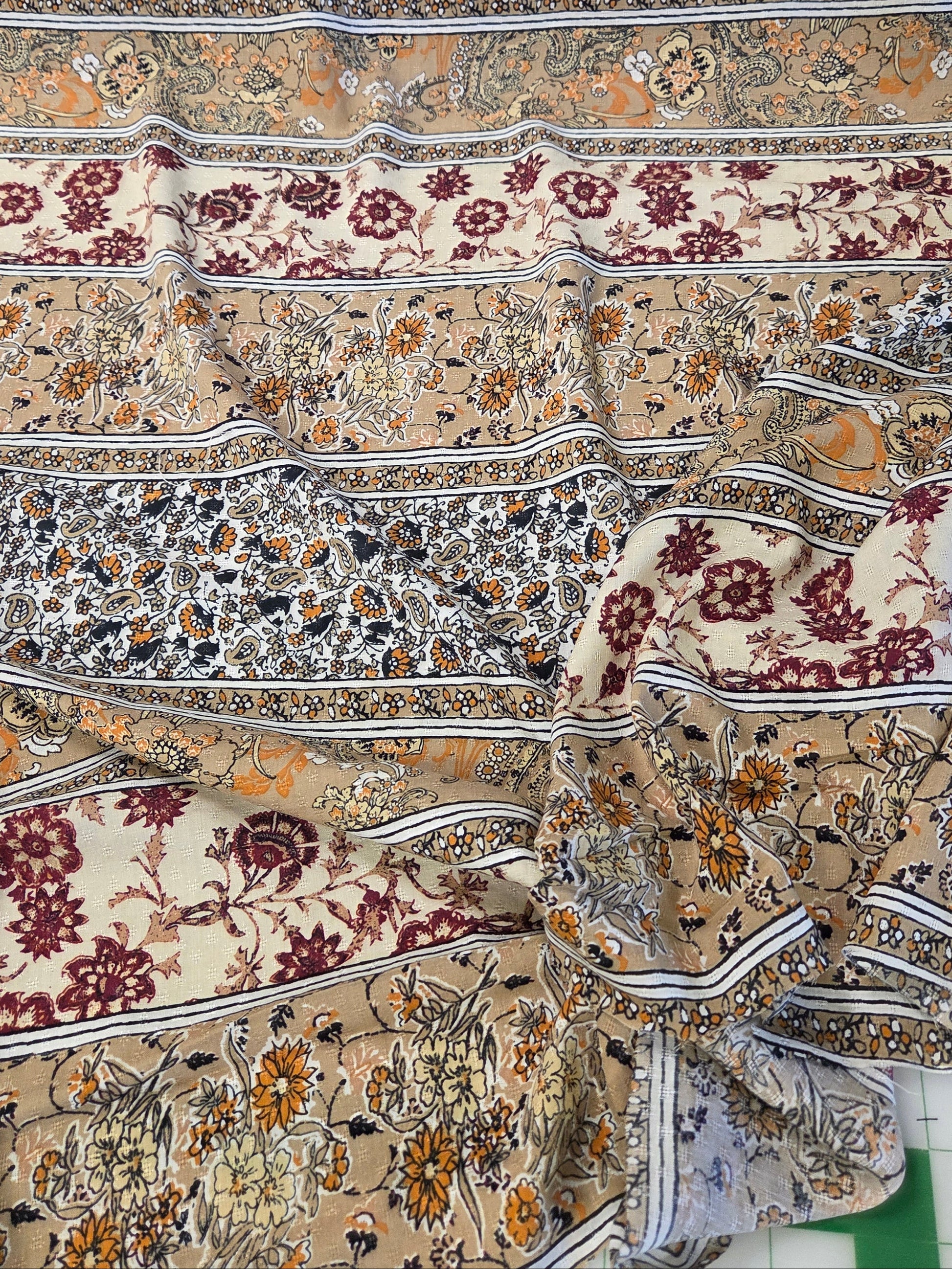 A draped piece of fabric with an intricate, multi-colored floral and ornamental pattern in beige, burgundy, orange, and gray, laid out on a green surface.