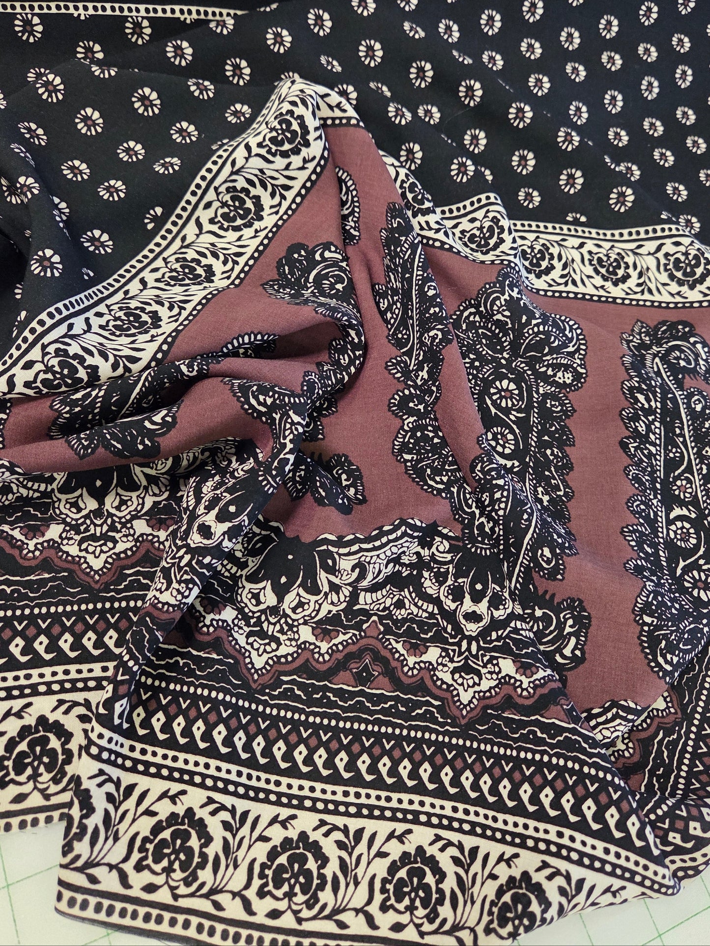 A folded and draped piece of fabric with a black base, featuring cream-colored floral and paisley patterns, and a dusty rose/mauve border with matching black and cream motifs.
