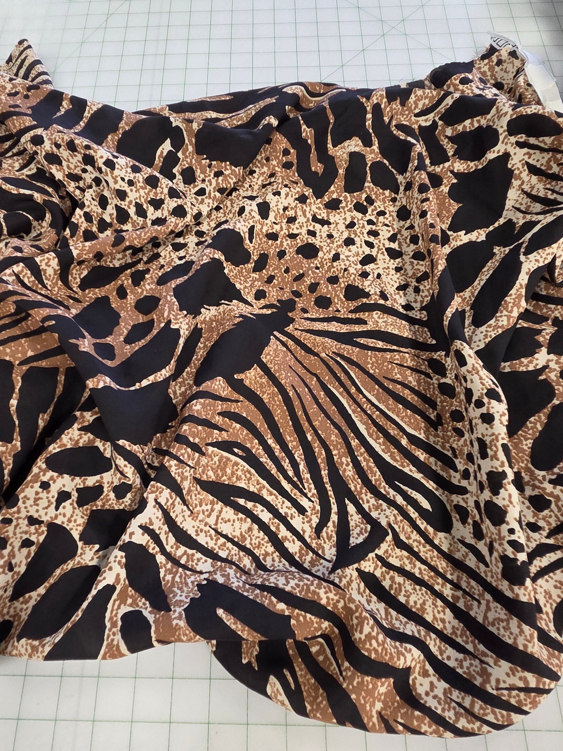 A leopard print fabric with a mix of black, brown, and beige animal spots and stripes, draped on a white gridded cutting mat in a sewing or tailoring room.