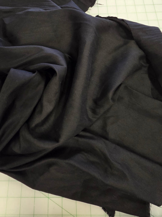 A large piece of black fabric, likely silk or satin, draped in soft folds on a white gridded cutting mat. The background shows a sewing room environment with a shelf, scissors, and other crafting supplies.