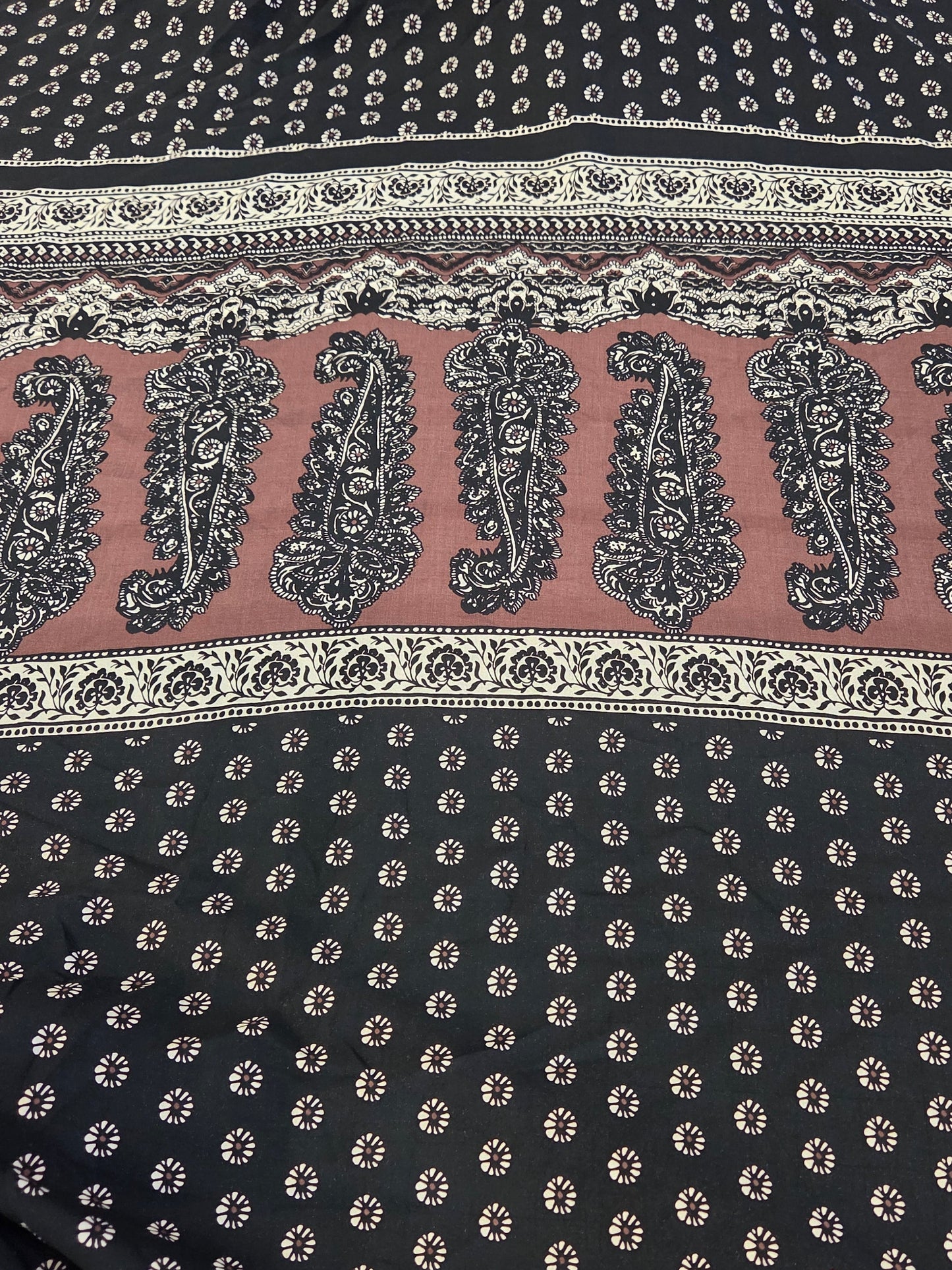 A close-up of a printed fabric with a black base, beige and white geometric borders, and a central dusty pink band featuring large black paisley motifs.
