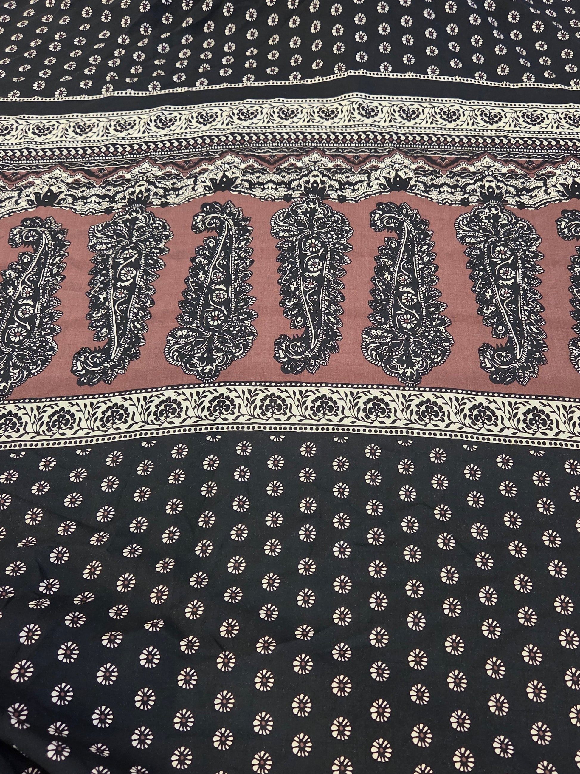 A close-up of a printed fabric with a black base, beige and white geometric borders, and a central dusty pink band featuring large black paisley motifs.
