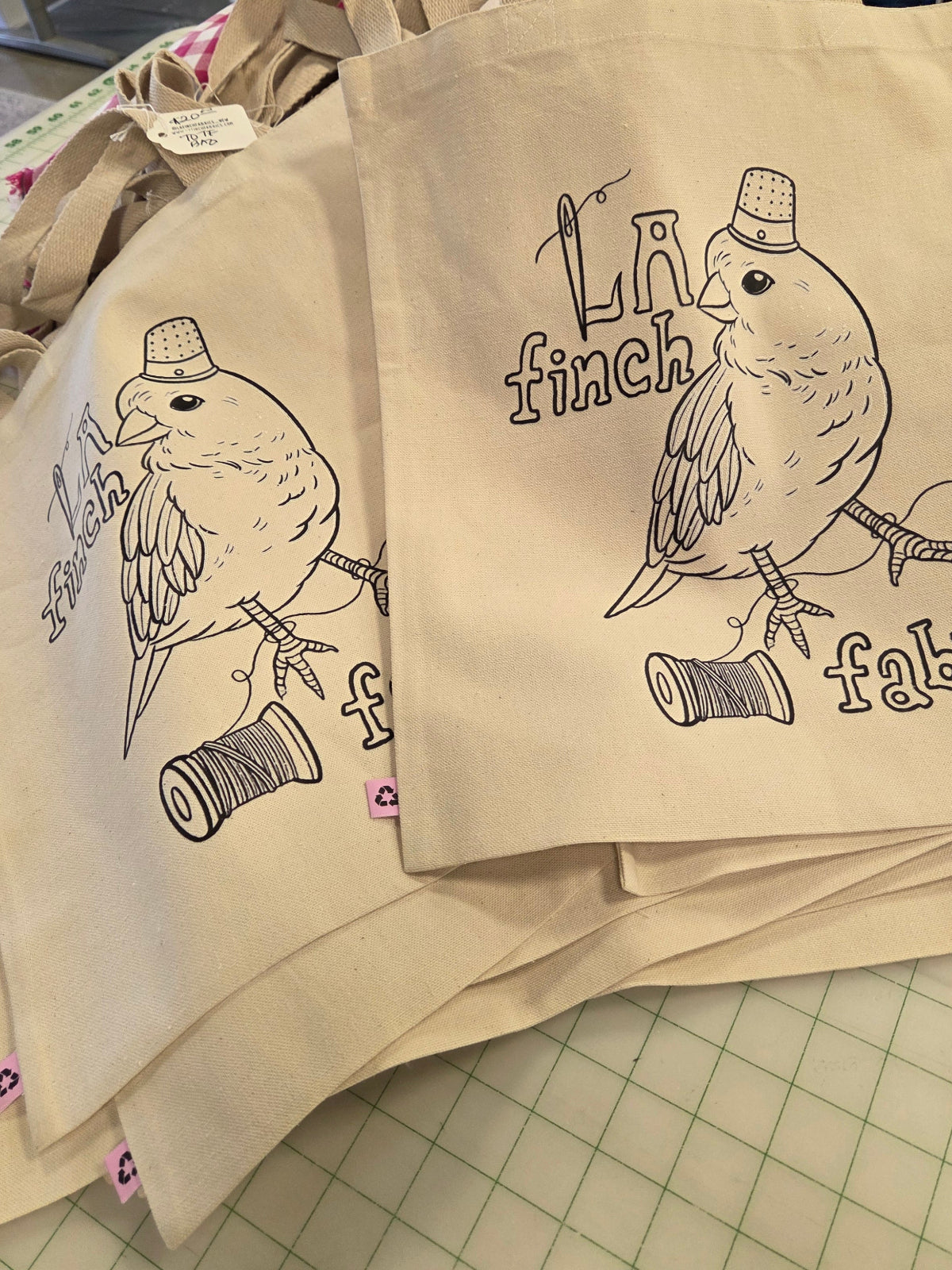 A stack of beige canvas tote bags with printed black line art of a bird wearing a top hat and the words 'La finch fabric,' photographed on a green grid cutting mat in a workshop setting.