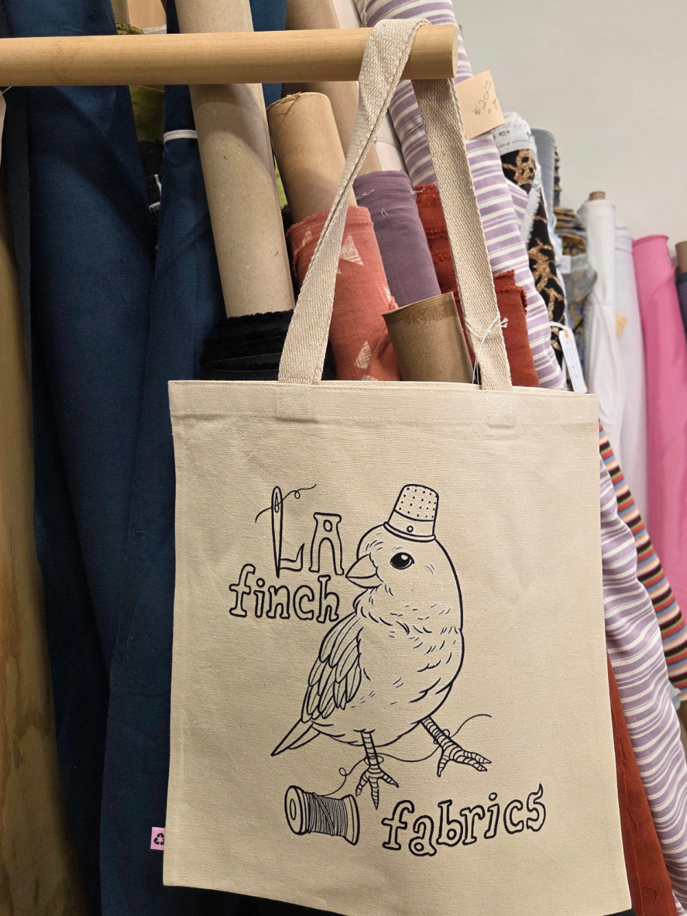A cream canvas tote bag with a black line art illustration of a finch wearing a top hat and the words 'finch fabrics', hanging from a wooden rod in front of a rack of colorful fabric rolls.