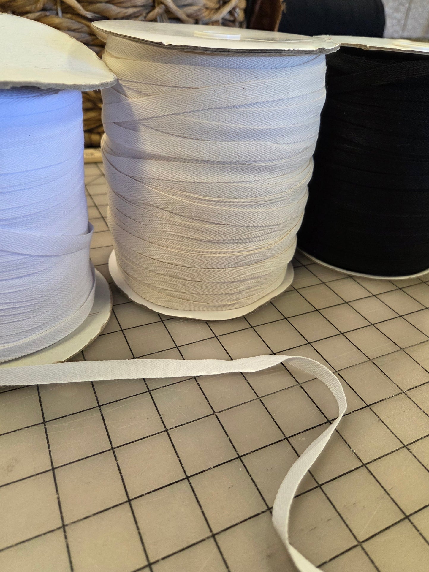 Three spools of ribbon or trim in white, off-white, and black, with a loose white strip laid on a gridded cutting mat in the foreground.