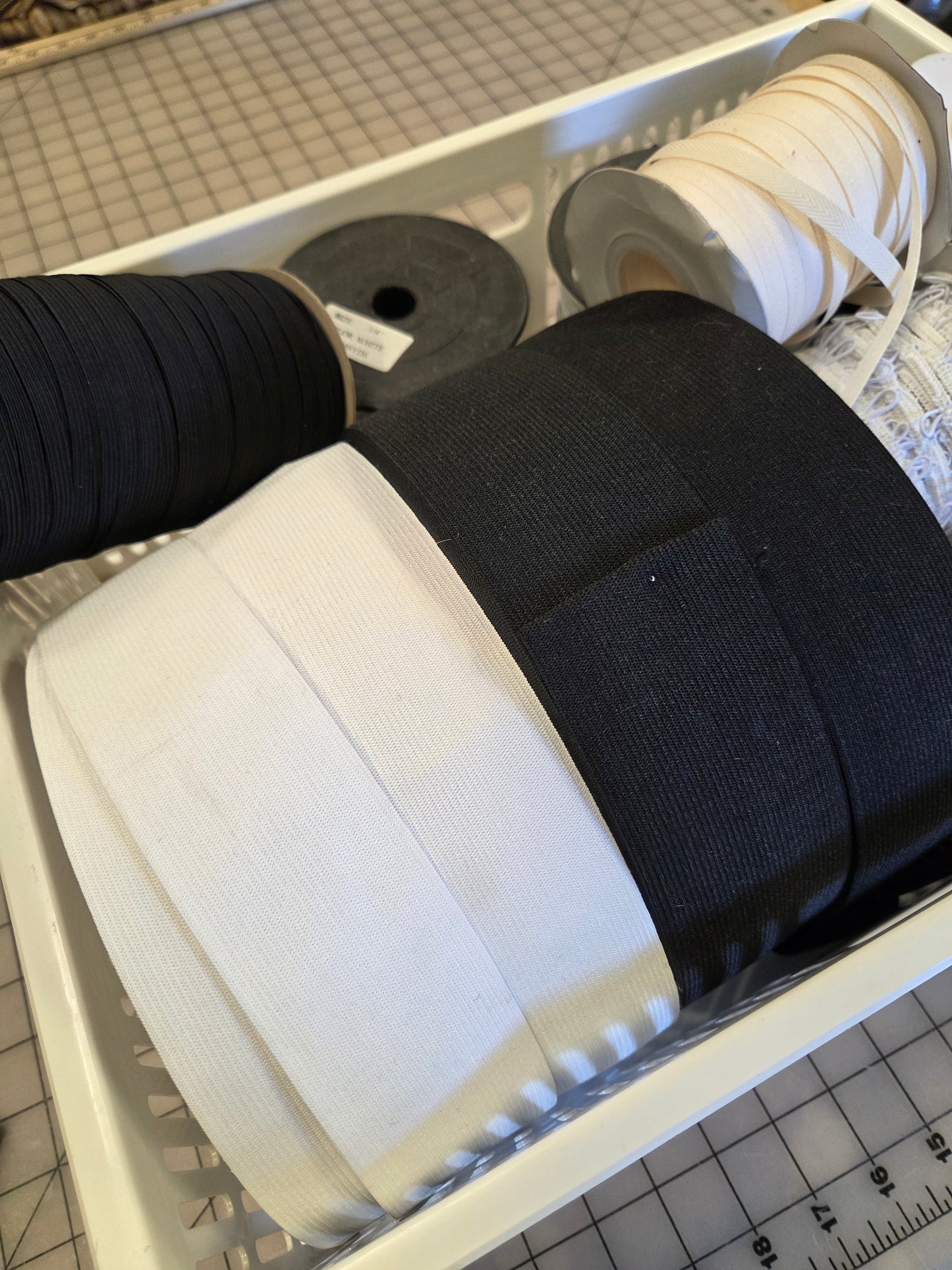 Spools of black and white elastic trim or ribbon spools arranged in a white plastic organizer tray on a gridded cutting mat.
