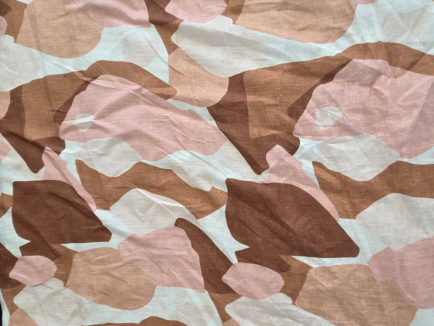A close-up of a crumpled fabric with an abstract camouflage pattern in shades of brown, dusty pink, beige, and off-white, laid flat on a dark surface with a wooden floor visible in the corner.