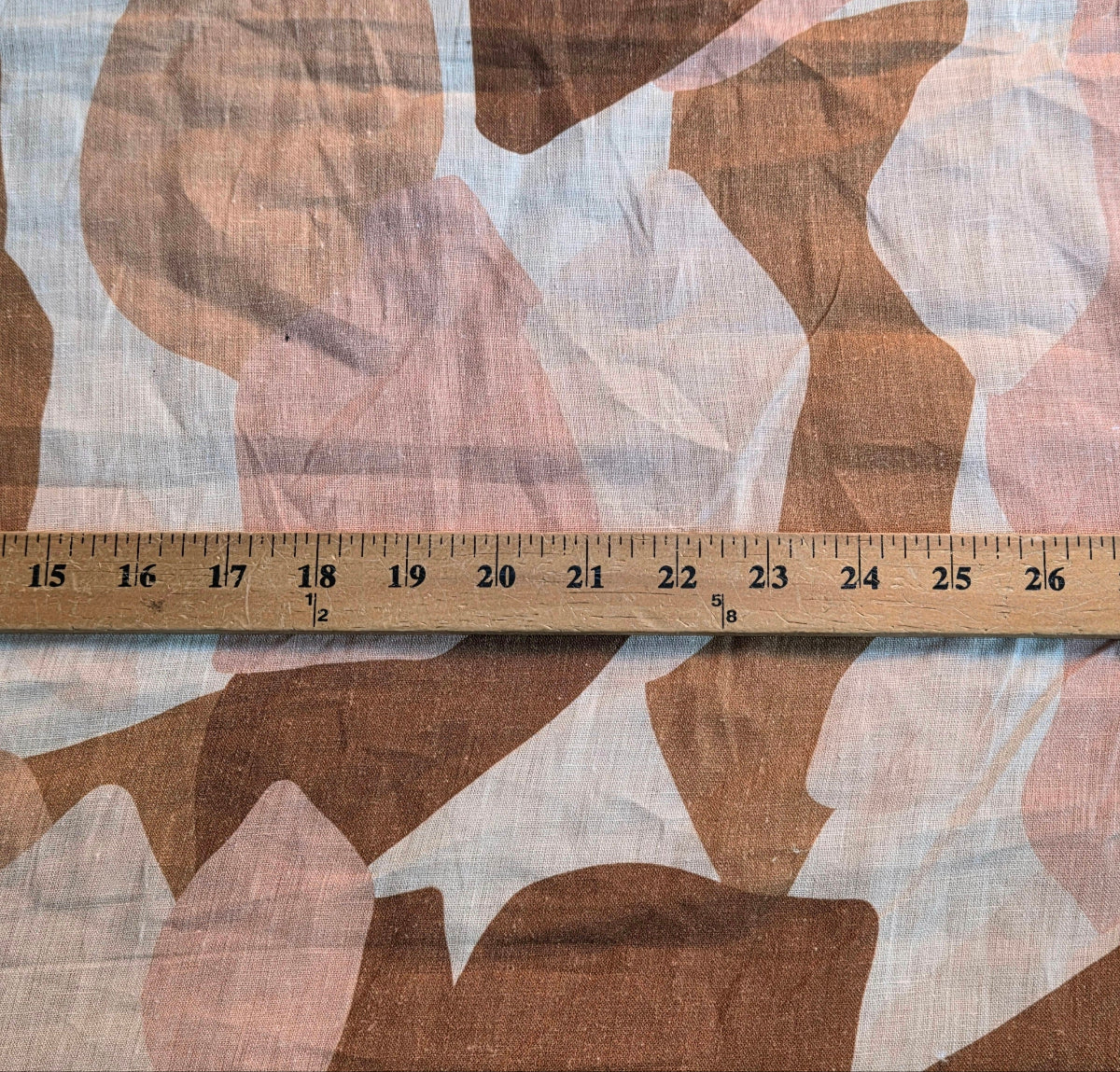 A close-up of a camouflage-patterned fabric with a wooden ruler laid horizontally across the center to show scale. The pattern consists of organic shapes in brown, dusty pink, and off-white.