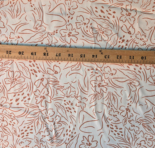 A close-up of a white fabric with a dusty rose or terracotta floral and leaf pattern, shown with a wooden ruler across the middle for scale.