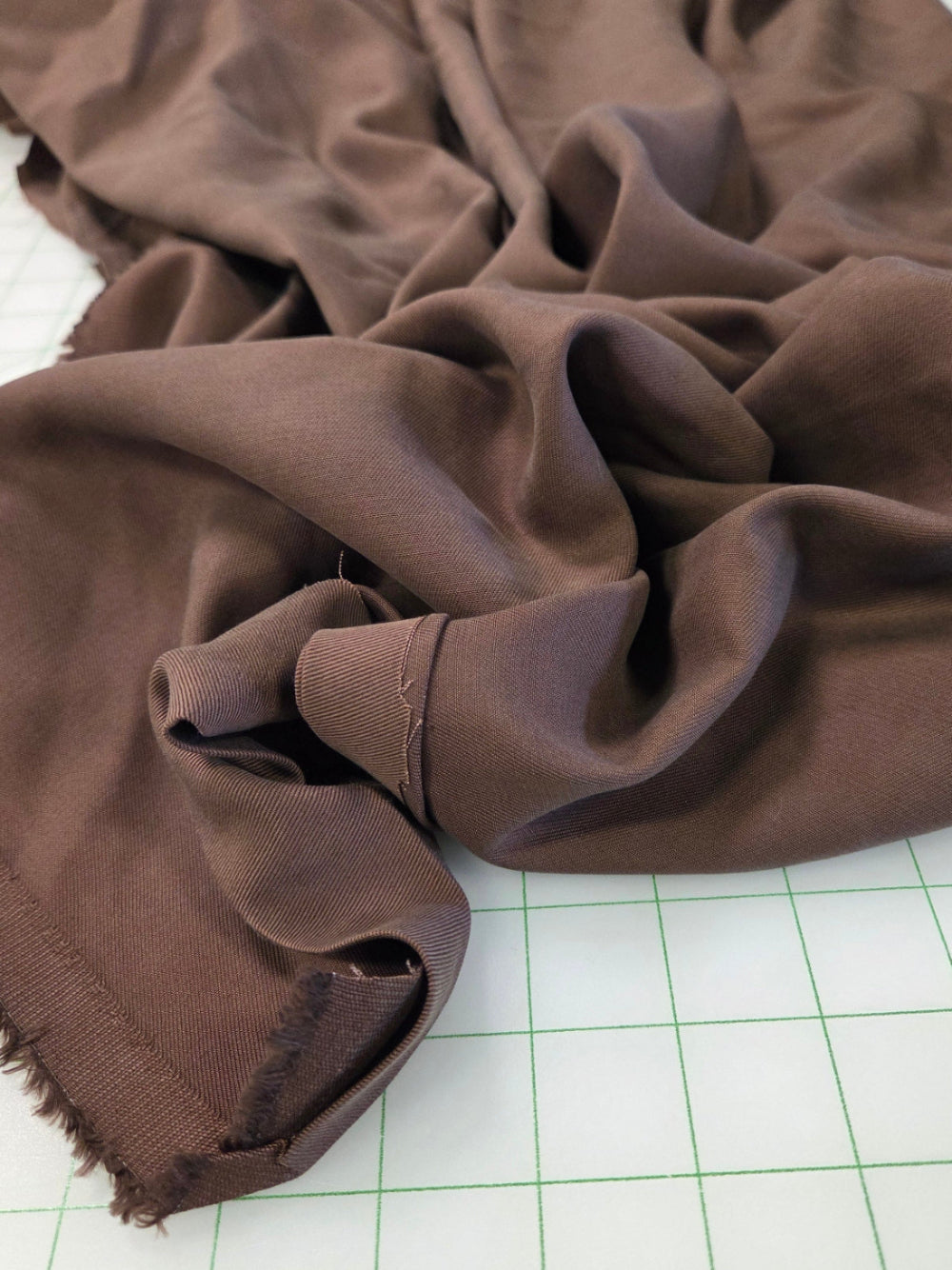 A close-up of draped brown fabric with a frayed edge, laid on a white cutting mat with green grid lines. Chocolate brown designer deadstock tencel twill woven solid for garment making