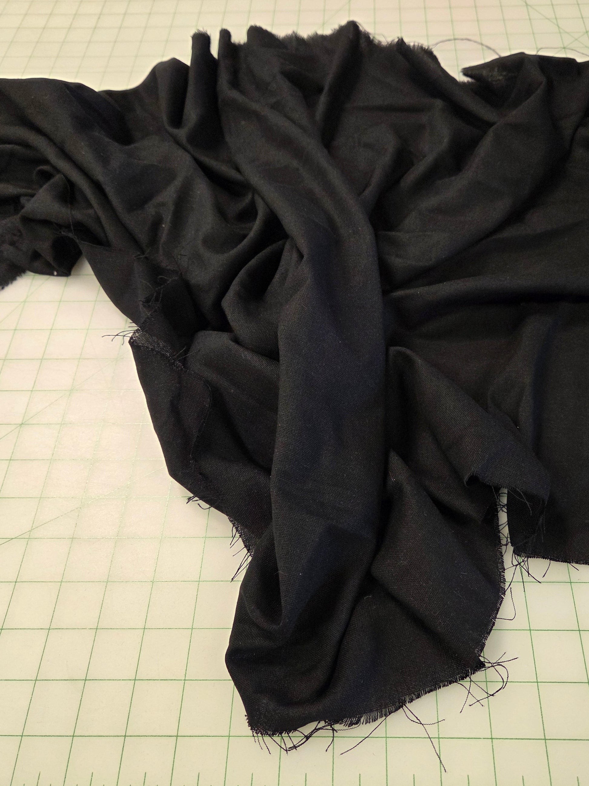 A large piece of draped black fabric with raw, frayed edges spread out on a green grid cutting mat, with a roll of black fabric visible in the top right corner.