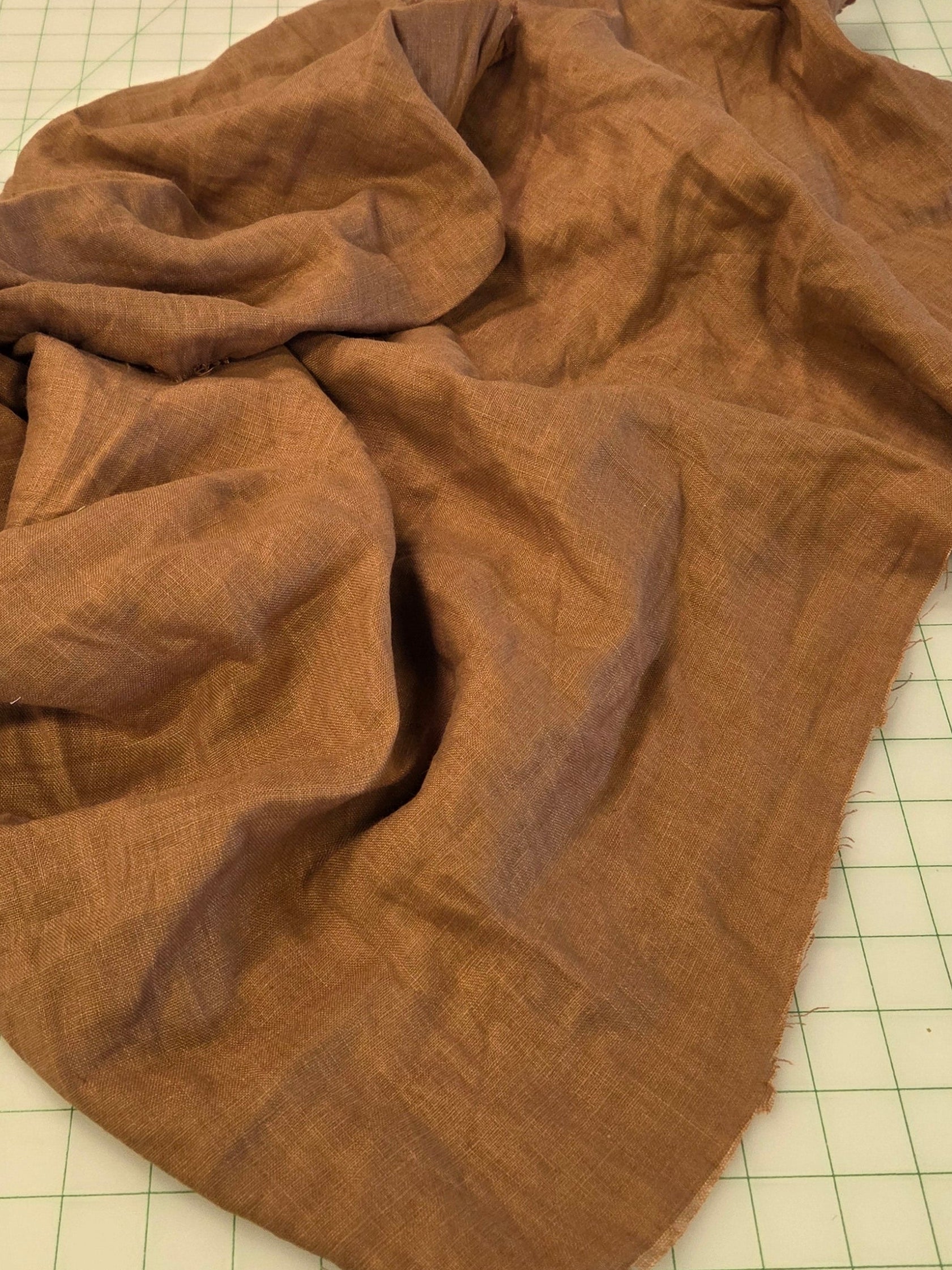 A crumpled brown linen fabric piece laid out on a green gridded cutting mat, with other fabrics and sewing supplies visible in the background.