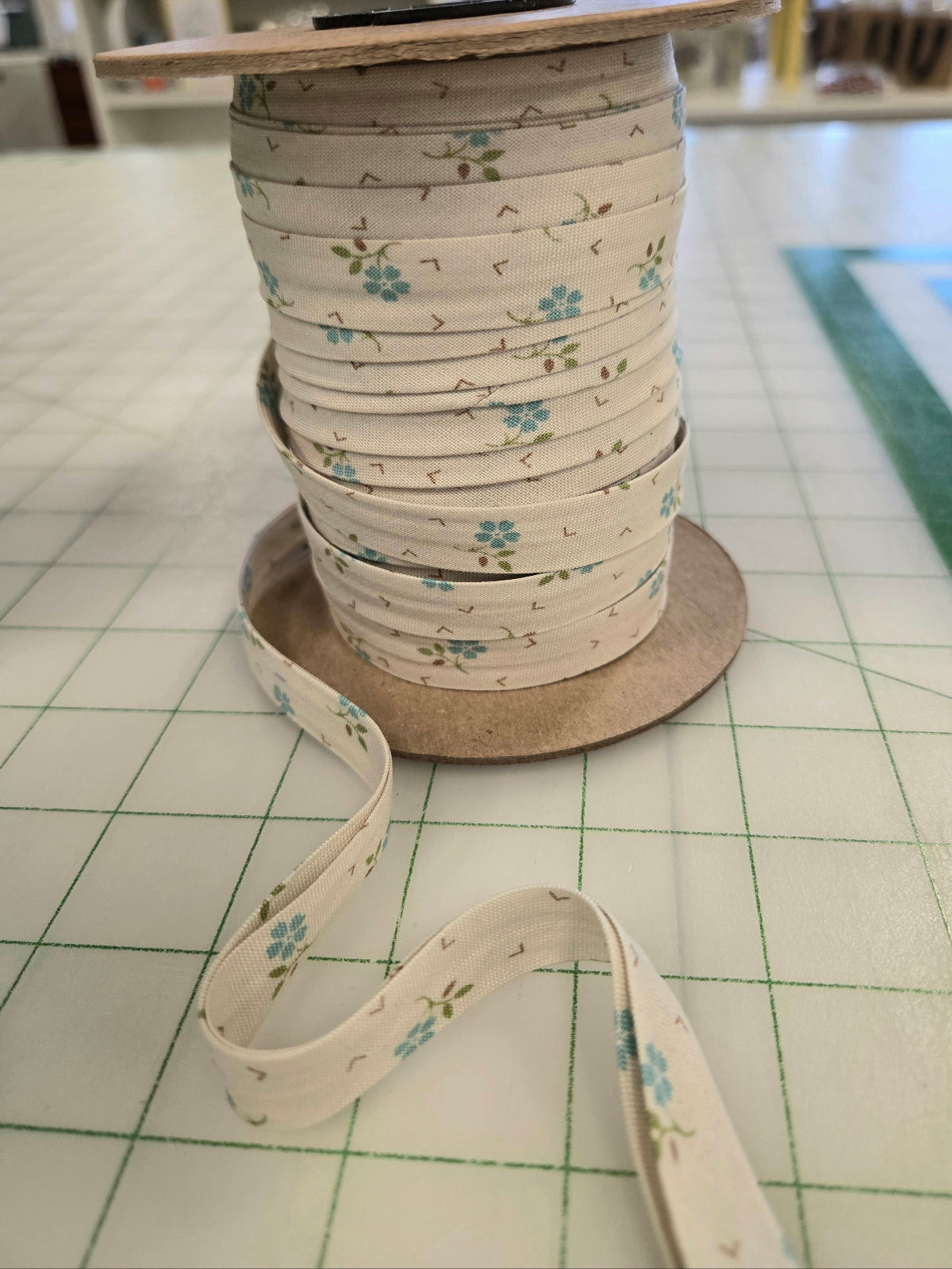 A spool of narrow floral ribbon with a cream base and small blue flowers with green stems, shown on a cutting mat in a craft room setting.