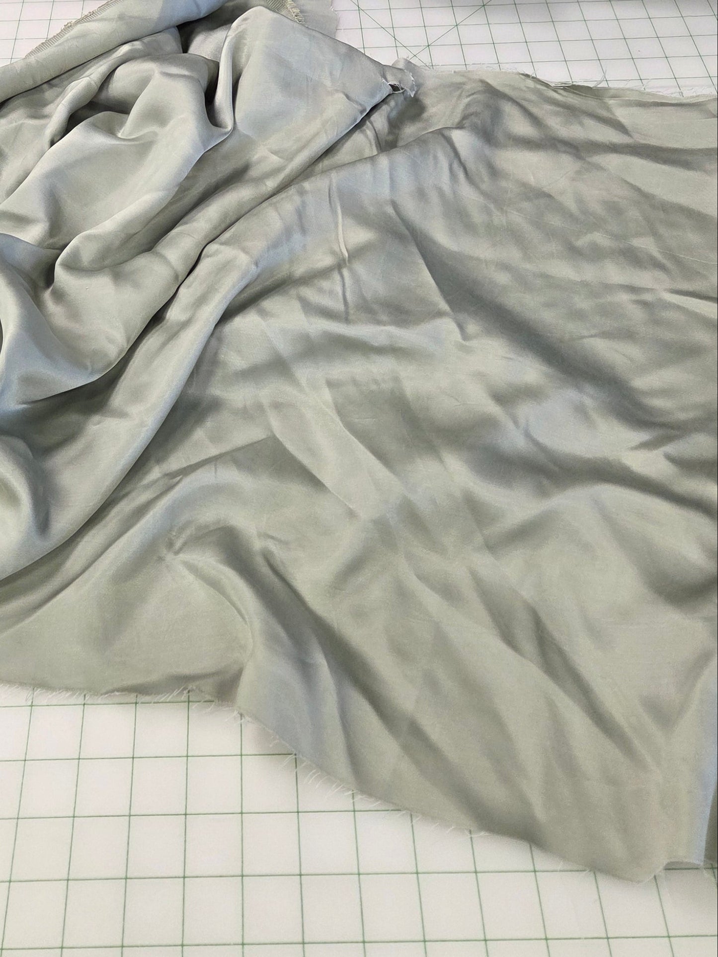 A light sage green fabric yardage is draped on a white gridded cutting mat, with a black fabric roll and a pile of black garments in the background inside what appears to be a sewing or fabric shop.