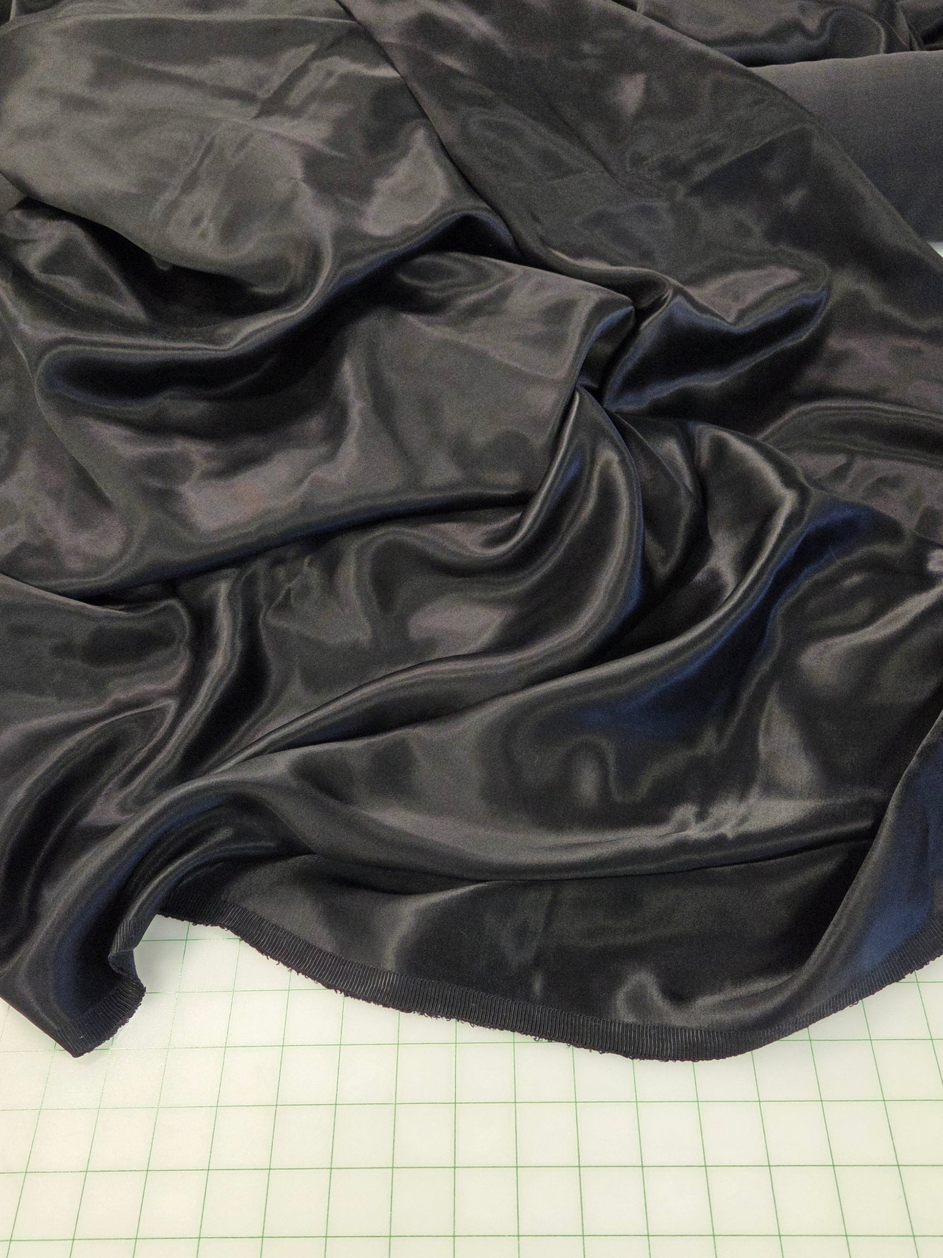 A draped piece of shiny black satin or silk fabric spread across a green gridded cutting mat, with a roll of matching black fabric visible in the background.