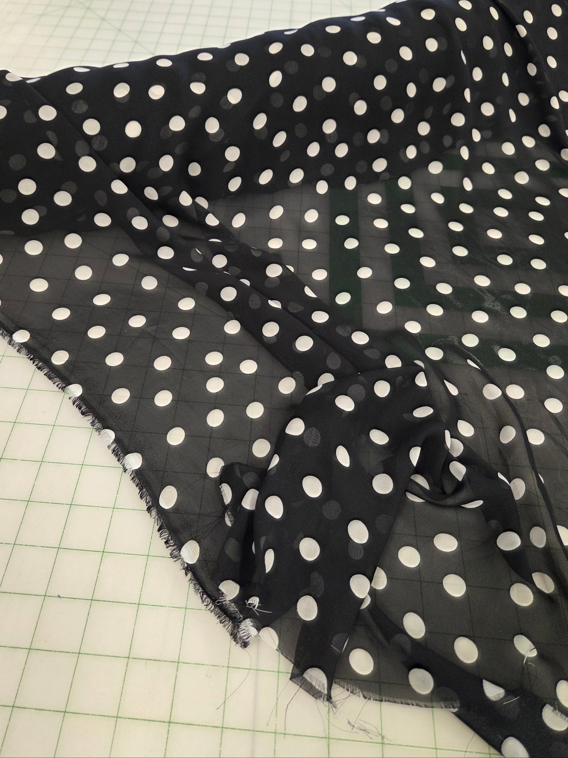 Black sheer fabric with a white polka dot pattern draped over a green grid cutting mat, with a sewing room visible in the background.