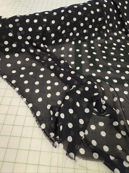 Black sheer fabric with a white polka dot pattern draped over a green grid cutting mat, with a sewing room visible in the background.