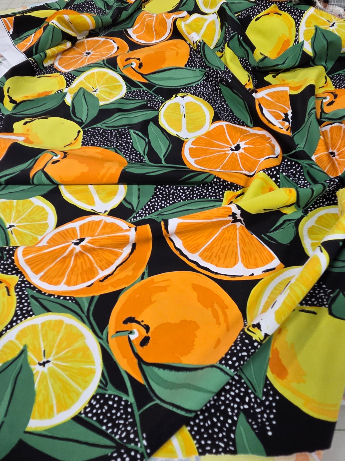 A close-up of a fabric or wallpaper pattern featuring a vibrant citrus design with orange slices, whole oranges, yellow lemons, and green leaves on a black background with white speckles.