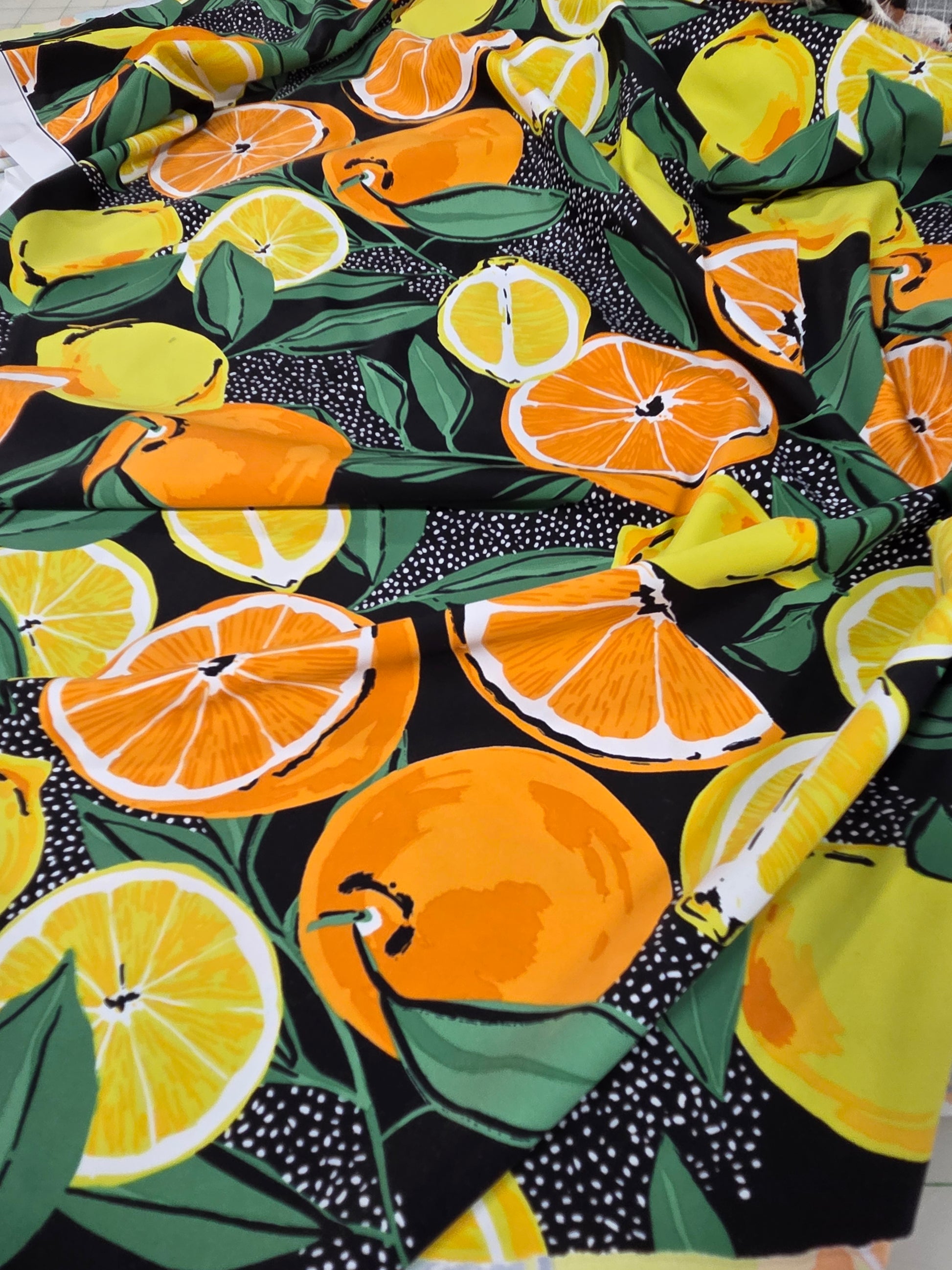 A close-up of a fabric or wallpaper pattern featuring a vibrant citrus design with orange slices, whole oranges, yellow lemons, and green leaves on a black background with white speckles.