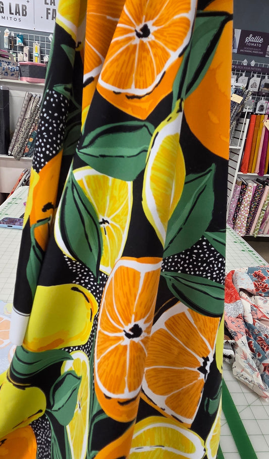 A close-up of a fabric yardage featuring a bold citrus pattern with orange slices, yellow lemons, green leaves, and black sections with white polka dots, displayed on a table inside a sewing supply store.