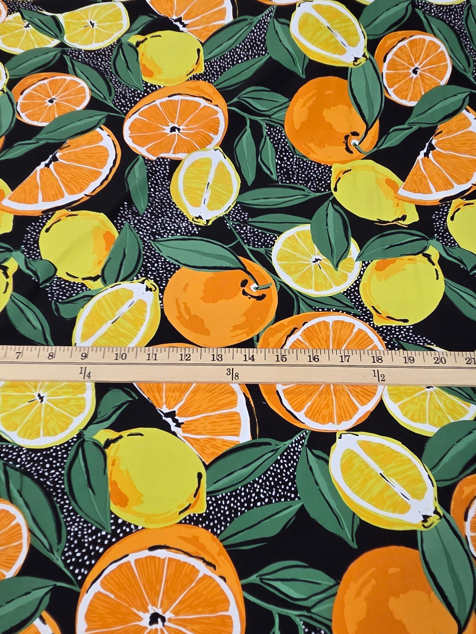 A close-up of a fabric or wallpaper pattern featuring a repeating print of lemons, oranges, and green leaves on a black background, with a wooden ruler placed horizontally to show scale.