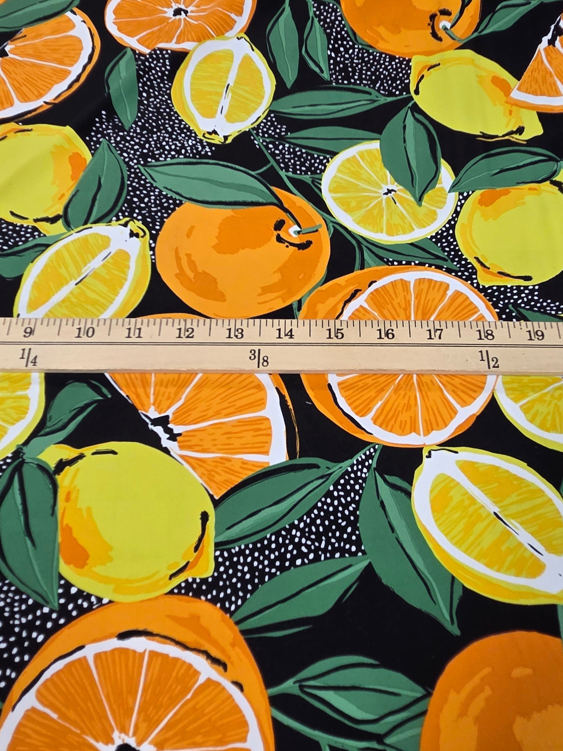A close-up of a fabric pattern featuring lemons, oranges, and green leaves on a black background, with a wooden ruler laid horizontally to show scale.