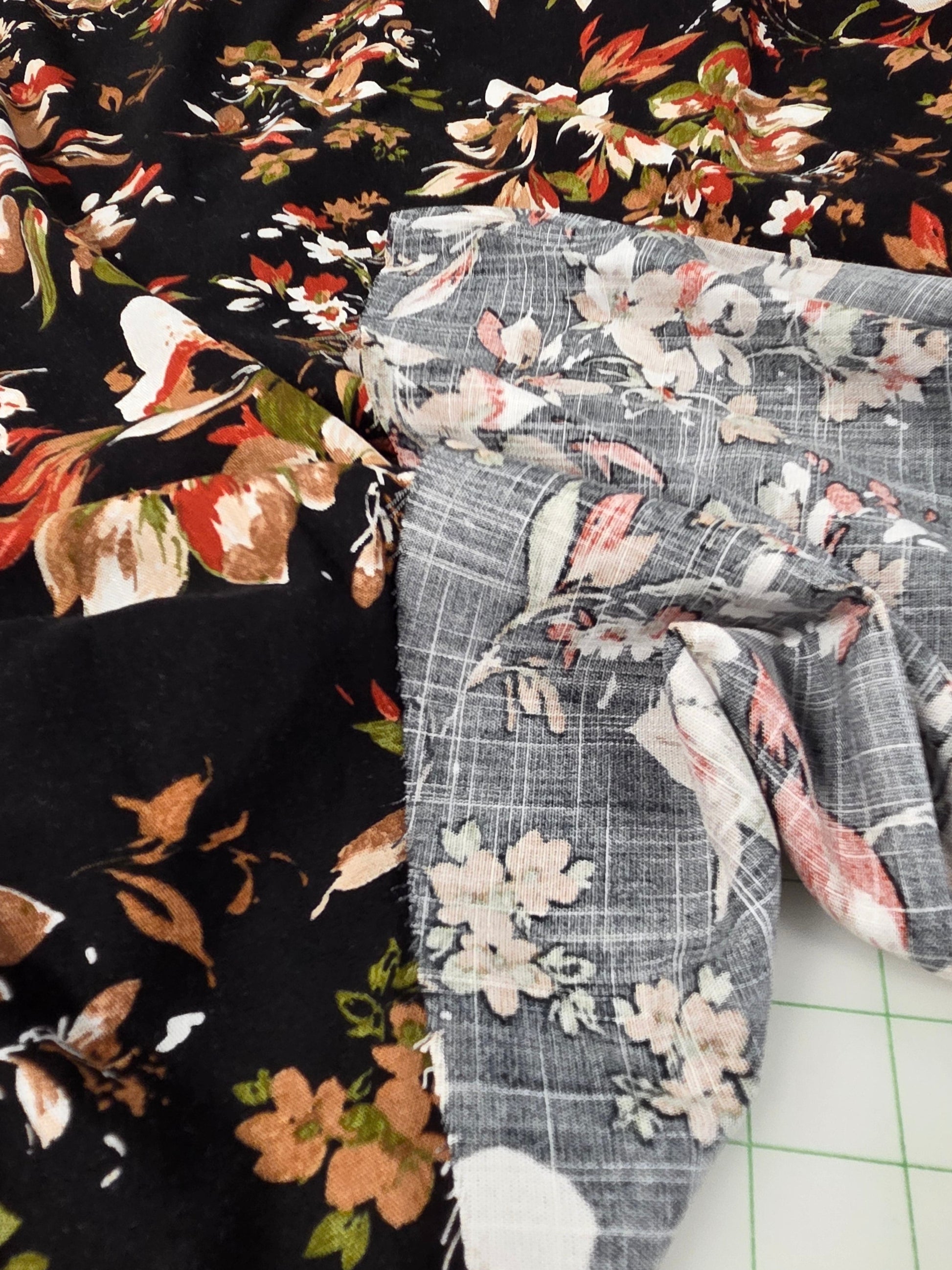Close-up of patterned fabric pieces laid flat, including a black floral textile with orange, white, and green floral motifs and a gray floral textile with white and pink flowers, on a white gridded cutting mat.